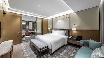 Habitación de hotel con una cama grande, una zona de estar con sofá y banco, un escritorio con teléfono, y un baño con espejo y lavamanos.