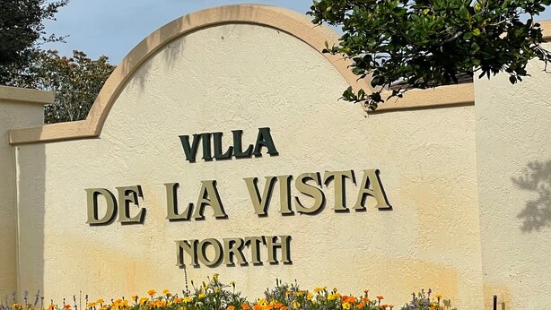 A sign for Villa De La Vista North with a decorative arch and a flower bed in front. - When Quality & Location Matter (The Villages)