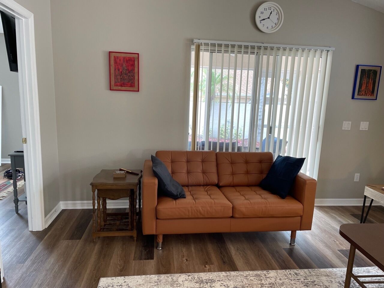 Ein Wohnzimmer mit einer Couch, einem kleinen Tisch, einer Wanduhr und einem Fenster mit Jalousien.