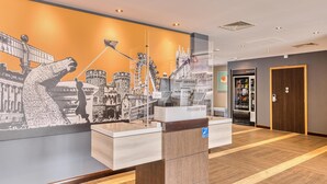 A hotel reception area with a large mural, a Premier Inn sign, a vending machine, and a wooden floor. - Premier Inn Bury St Edmunds North - A14 (Bury St Edmunds)