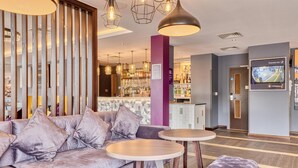 A modern lobby with a sofa, round tables, a bar area, and a television screen. - Premier Inn Bury St Edmunds North - A14 (Bury St Edmunds)