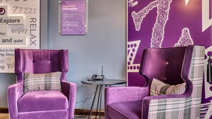 A room with purple upholstered chairs, a small round table, and a wall with decorative patterns and text. - Premier Inn Bury St Edmunds North - A14 (Bury St Edmunds)