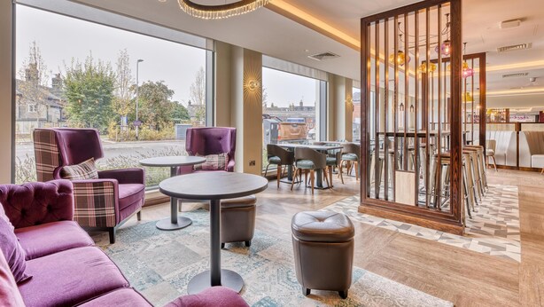 A modern hotel lobby with large windows, plush seating, a round table, and a decorative partition. - Premier Inn Bury St Edmunds North - A14 (Bury St Edmunds)