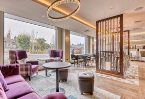 A modern hotel lobby with large windows, plush seating, a round table, and a decorative partition.