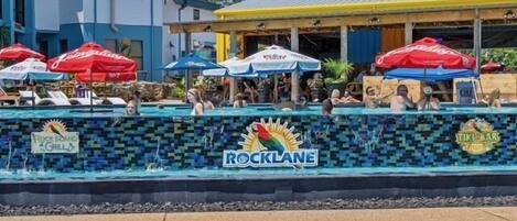 A pool area with lounge chairs and umbrellas, a sign for 'Rocklane', and a 'Tiki Bar' sign.