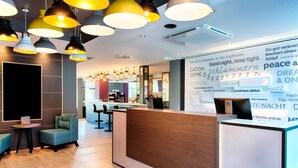 Un espace d’accueil d’hôtel avec un comptoir de réception en bois, plusieurs lampes suspendues et un coin salon avec des canapés.
