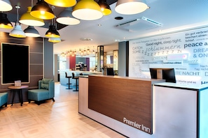 A hotel reception area with a wooden reception desk, multiple hanging pendant lights, and a seating area with sofas. - Premier Inn Lübeck City Stadtgraben (Lubeck)