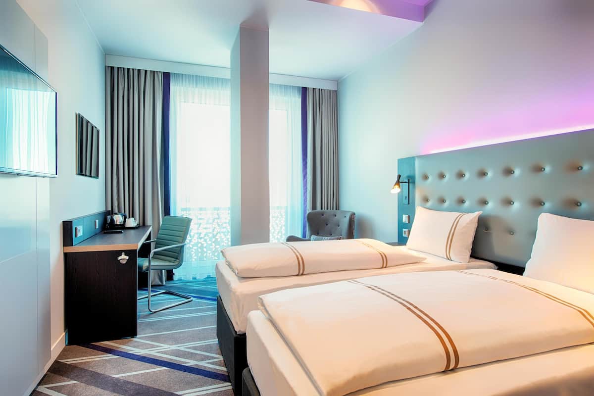 A hotel room with two beds, a desk, a chair, and a view of the outdoors.