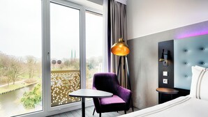 Une chambre d’hôtel dotée d’une grande fenêtre, d’un fauteuil violet, d’une table ronde, d’un lit et d’une tête de lit moderne.