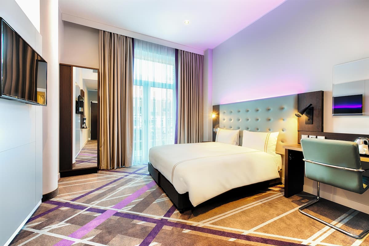 A modern hotel room with a large bed, a seating area, a television, and a view of the city.