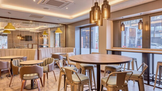A modern restaurant interior with round tables, cushioned seating, pendant lighting, and large windows offering city views. - Premier Inn London Uxbridge Town Centre (Uxbridge)