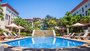 A swimming pool with lounge chairs and umbrellas, surrounded by palm trees and a stone wall.