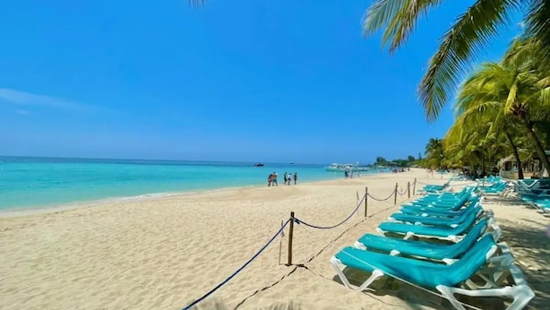 A beach with clear turquoise water, a sandy shore, and rows of blue lounge chairs.