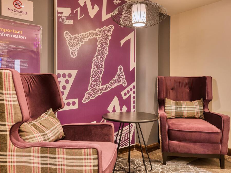 A hotel lobby with a 'No Smoking' sign, a 'Important Information' poster, a decorative wall with a large 'I' design, and two plush armchairs with plaid cushions.