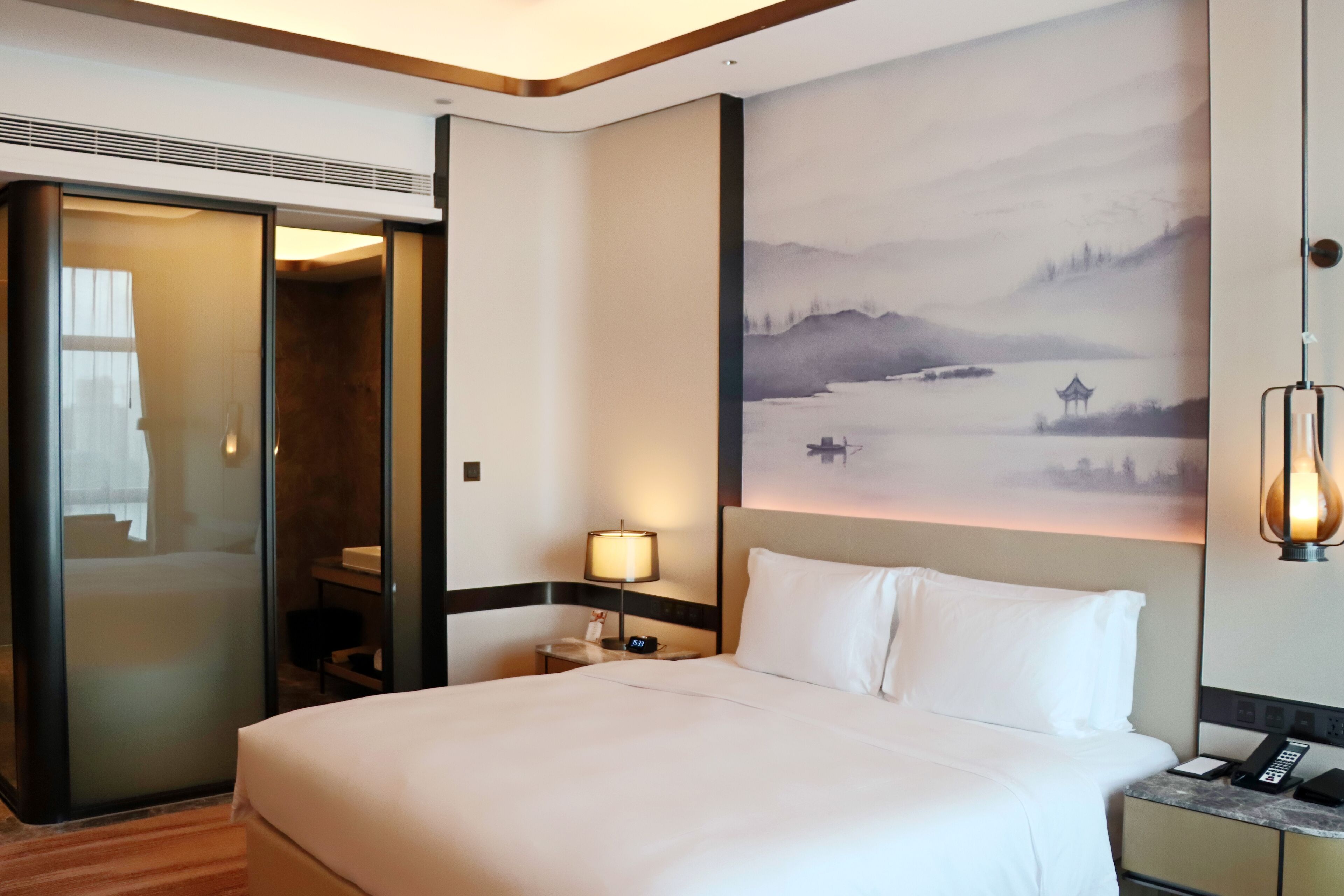 A hotel room with a large bed, a nightstand with a lamp, a painting on the wall, and a sliding door leading to a bathroom.