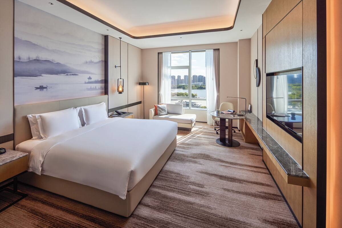 A hotel room with a large bed, a desk, a chair, a TV, and a view of a cityscape through the window.
