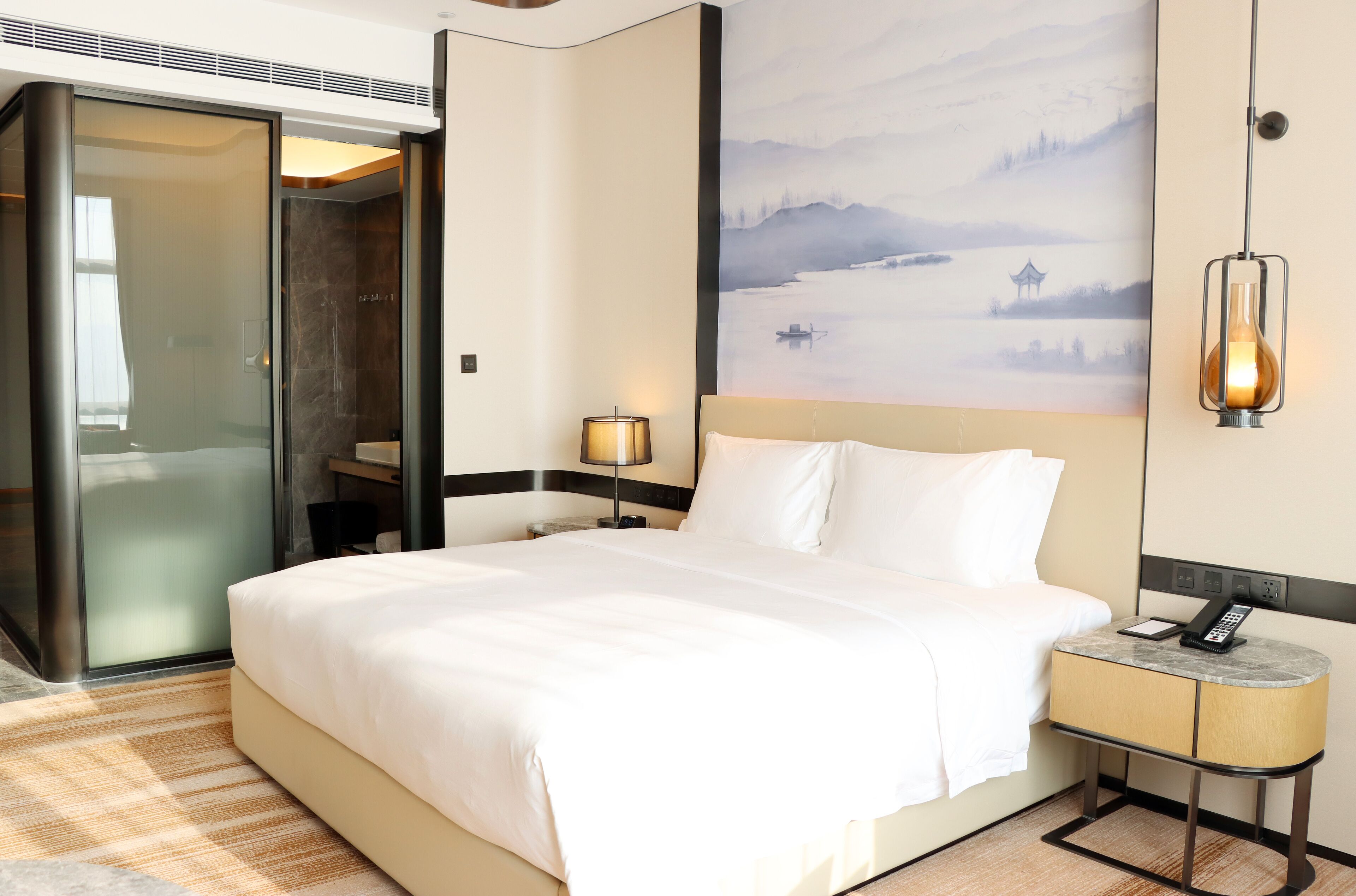 Quarto de hotel com uma cama grande, uma mesinha de cabeceira com abajur, um quadro de uma paisagem calma e uma porta deslizante para o banheiro.