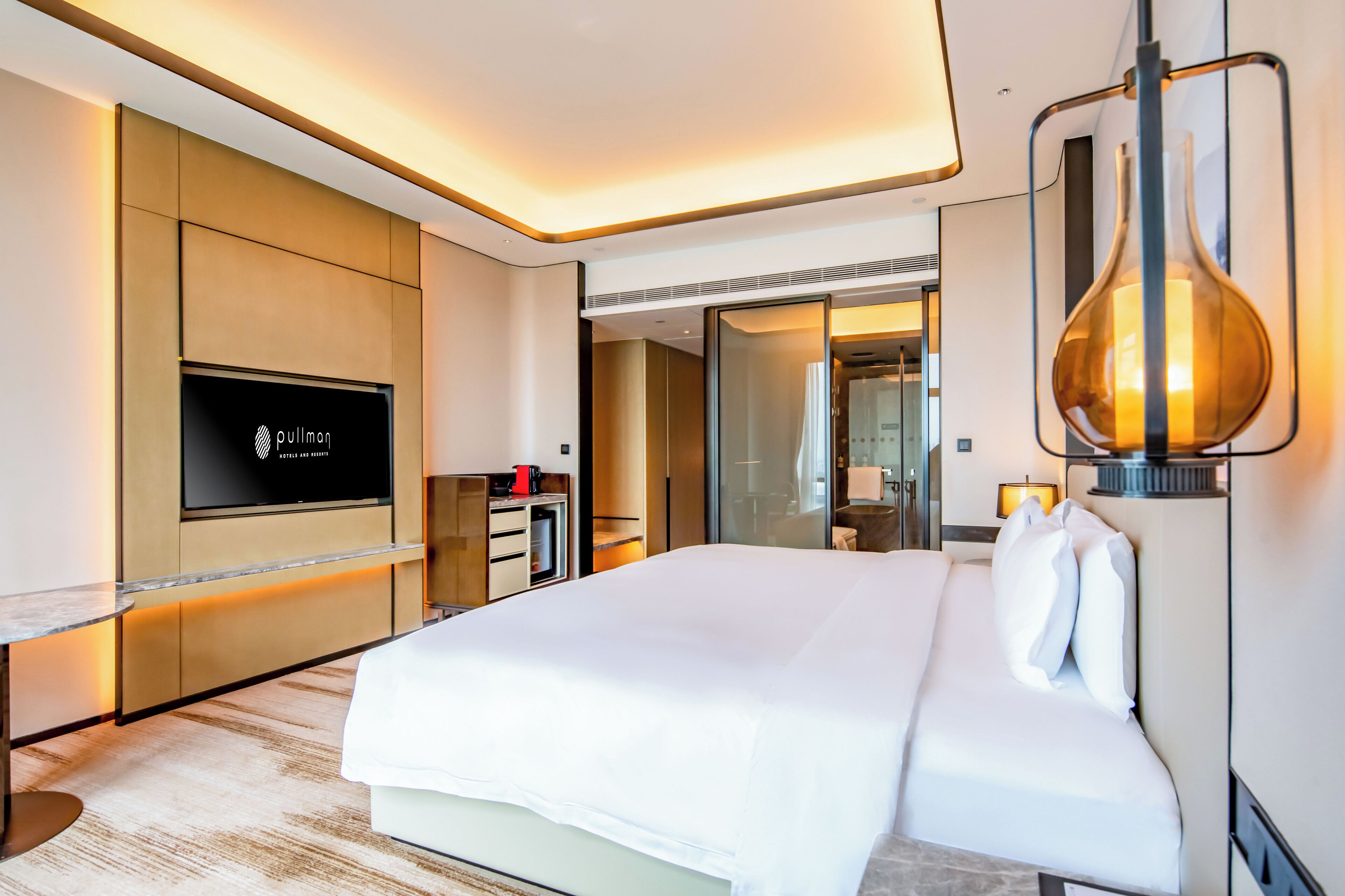 Una habitación de hotel moderna con una cama grande, un televisor de pantalla plana, un minibar y un baño con paredes de vidrio.
