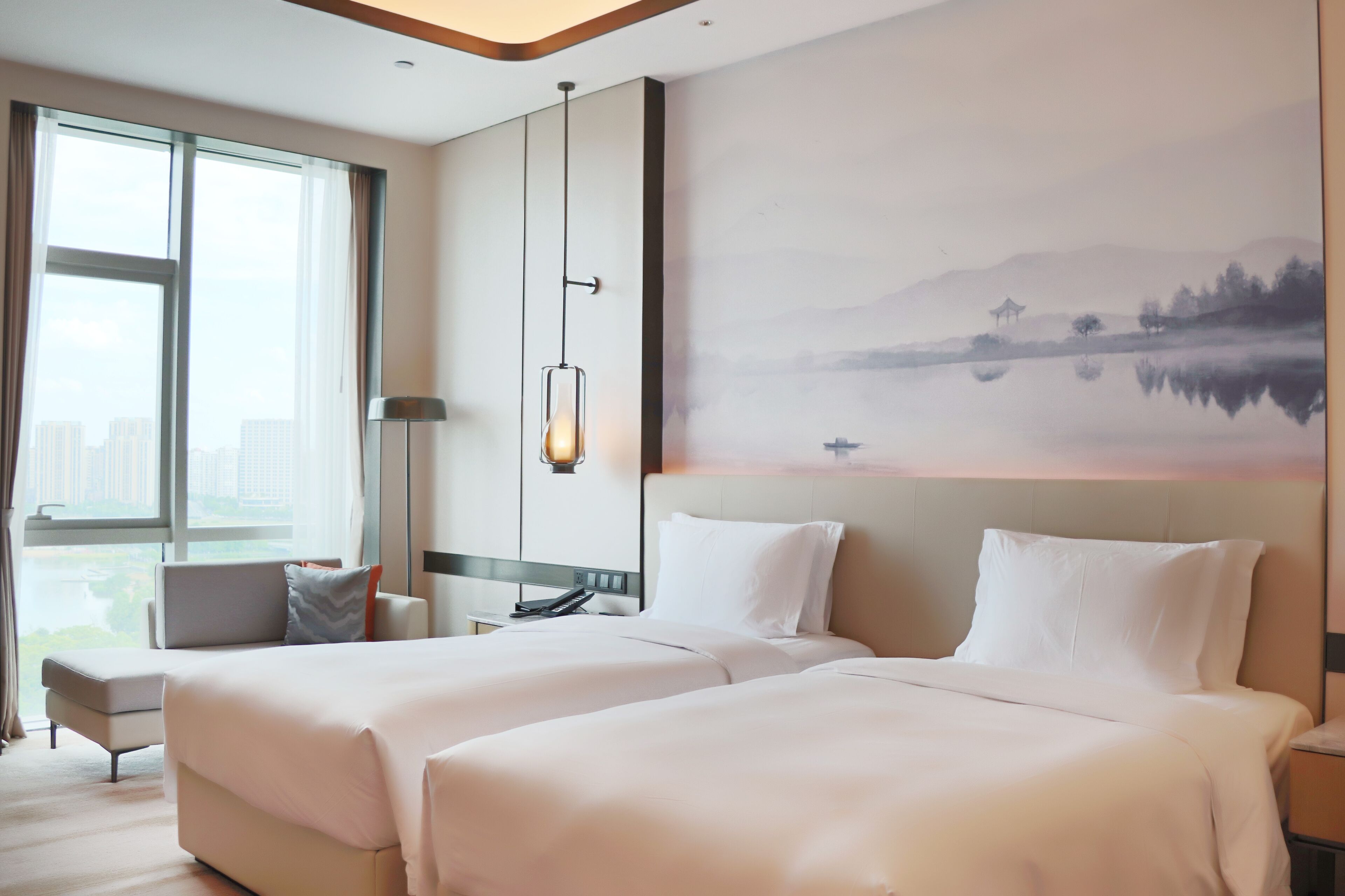 Habitación de hotel con dos camas, un ventanal grande, un cuadro de un paisaje y una lámpara.