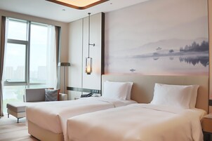 Habitación de hotel con dos camas, un ventanal grande, un cuadro de un paisaje y una lámpara.