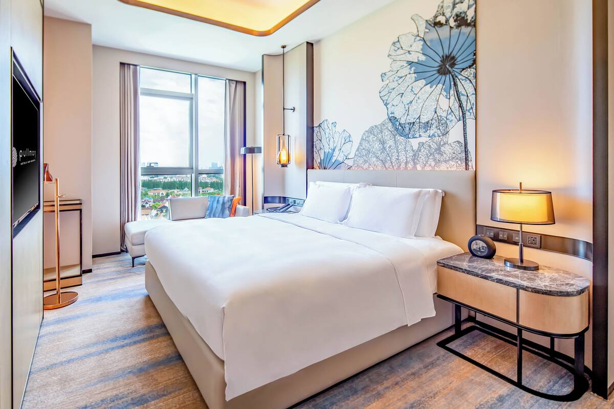 A hotel room with a large bed, a desk with a lamp, a TV, and a view of the city.