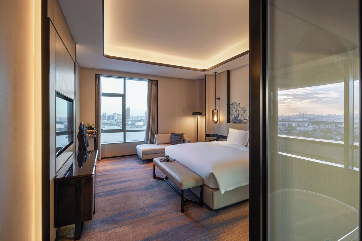 A modern hotel room with a large bed, a seating area, a desk, and a city view.