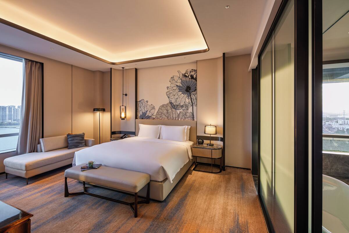 A modern hotel room with a large bed, a sofa, a bench, a bedside table with a lamp, and a view of the city through the window.