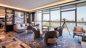 A modern hotel lobby with a large window overlooking a body of water and cityscape. There are comfortable seating areas, a large bookshelf, and a tripod with a camera. - Pullman Suzhou Taicang  (Suzhou)