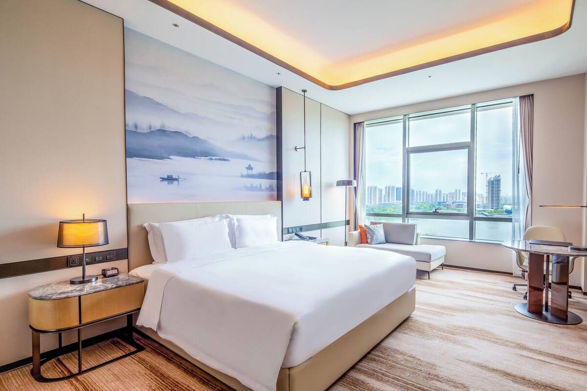 A hotel room with a large bed, a desk, a chair, and a view of a cityscape through the window.