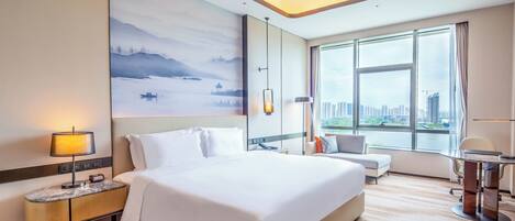 A hotel room with a large bed, a desk, a chair, and a view of a cityscape through the window.