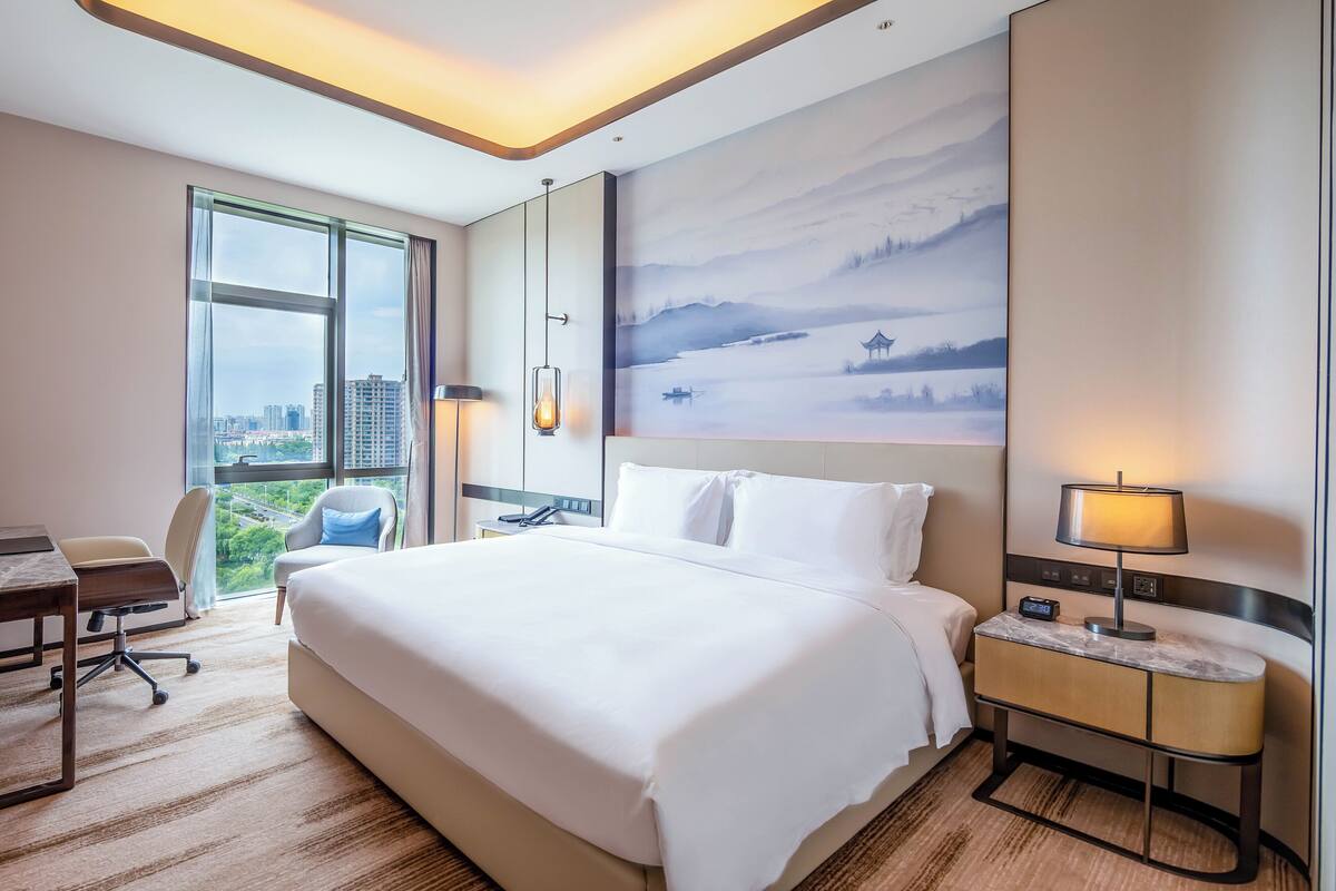 A hotel room with a large bed, a desk, a chair, a nightstand, and a painting on the wall.