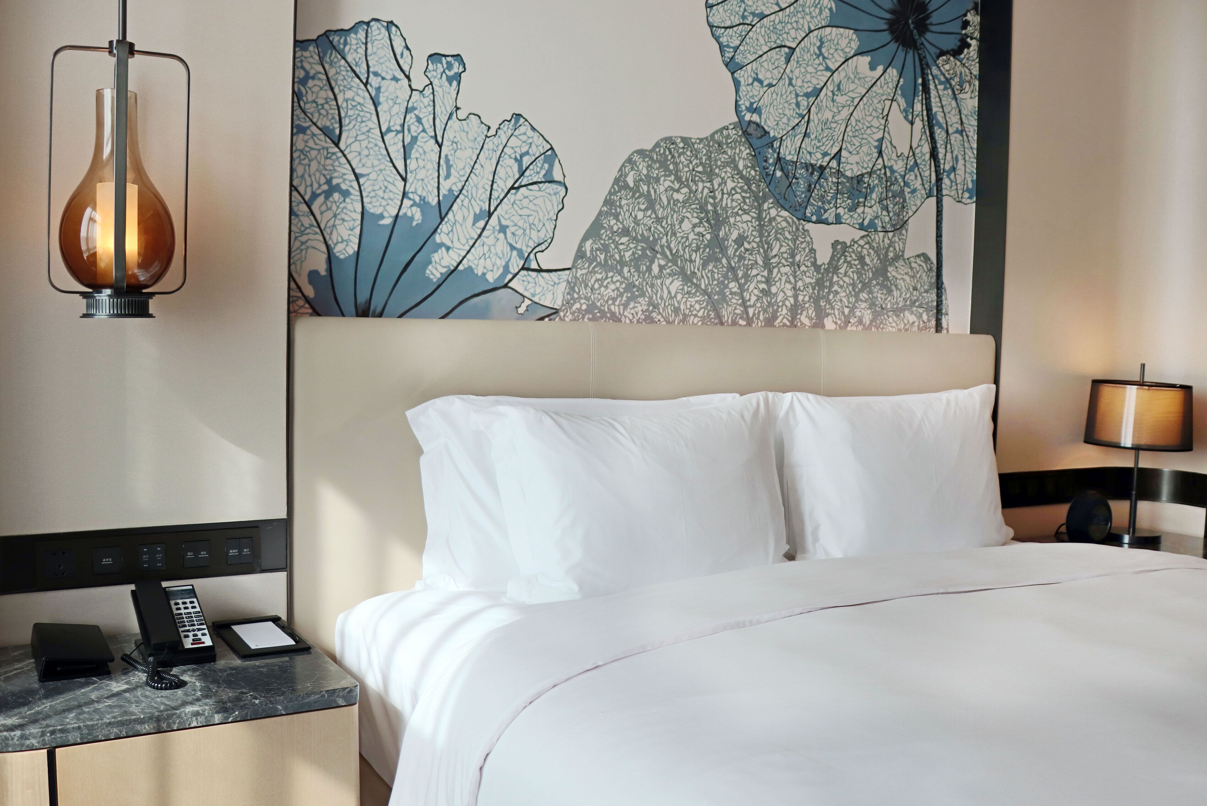 A hotel room with a bed, a nightstand with a phone, a lamp, and a wall art piece.