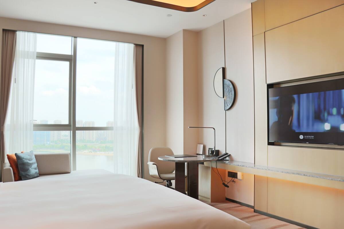 A hotel room with a large bed, a desk with a chair, a TV mounted on the wall, and a window with curtains.
