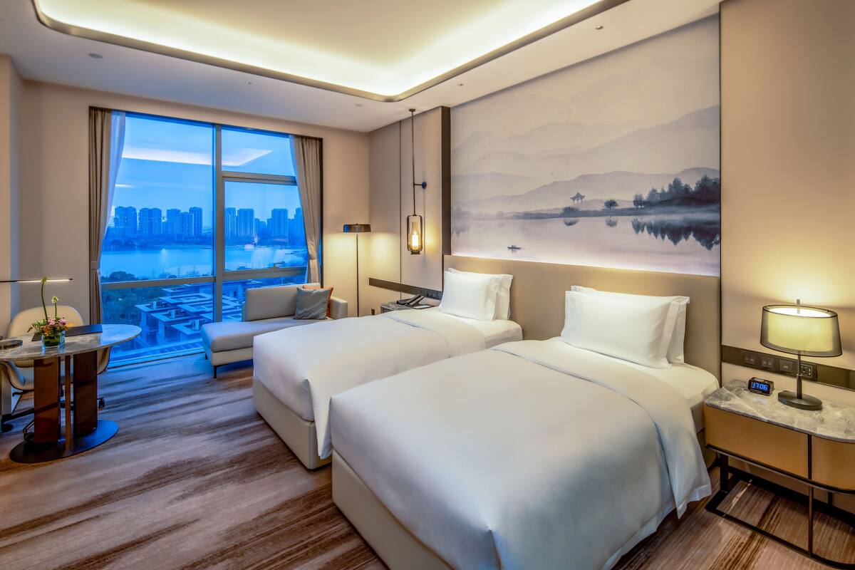 A hotel room with two beds, a desk, a chair, and a large window offering a city view.