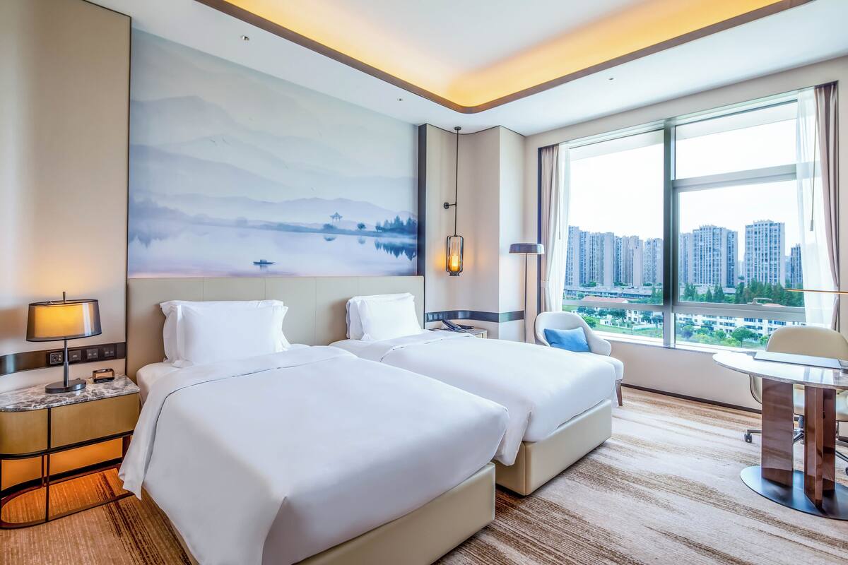 A hotel room with two beds, a desk, a chair, a large window with a city view, and a painting on the wall.