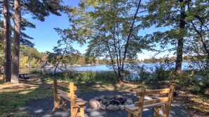 A wooden bench and picnic table set up near a lake with a fire pit in the center.