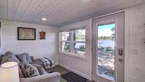 A cozy room with a wooden ceiling, a grey sofa with patterned cushions, a framed picture on the wall, and a large window offering a view of a lake.