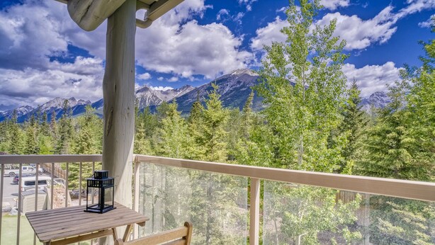 Un balcone con tavolo e sedia in legno, affacciato su un paesaggio montuoso con alberi e cielo sereno.