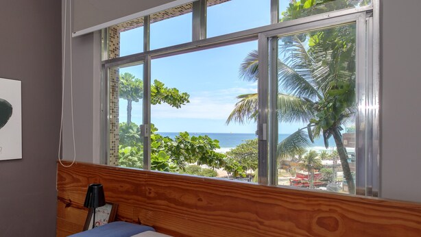 A bedroom with a large window overlooking a beach and palm trees. - Pepezinho Ocean View (Rio de Janeiro)