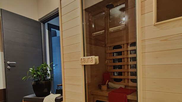 A sauna room with wooden walls and a glass door, a small table with a plant, and a red towel on the bench. - Casa nel Bosco con Sauna (Cervasca)