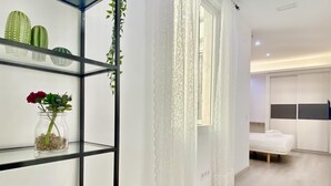 Et moderne hotelværelse med en glasreol med dekorative genstande, en seng med hvid madras og puder, og et stort vindue med lette gardiner.