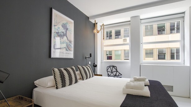Une chambre moderne avec un grand lit, un coussin aux motifs noir et blanc, une œuvre d’art encadrée, une chaise noire et une fenêtre donnant sur des immeubles.