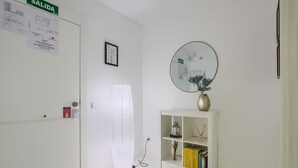 Un couloir avec une porte blanche portant l’inscription « SORTIE », une étagère blanche avec des livres et des objets décoratifs, et un miroir rond accroché au mur.