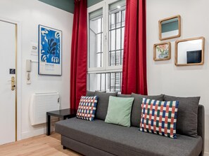 A grey sofa with patterned pillows, a white door, a window with red curtains, and a framed artwork on the wall.