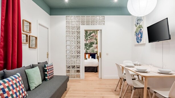 Een moderne woonkamer met een grijze bank, kleurrijke kussens, een eethoek met een witte tafel en stoelen, en een aan de muur bevestigde televisie.