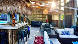 An outdoor patio with a thatched roof, wooden deck, bar area, seating, and a pool in the background. - 4 Decks, Private Pool, Swim-up Tiki Bar, Hot Tub (North Captiva)