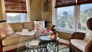 A cozy living room with a sofa, a wicker chair, a glass coffee table, and a view of palm trees through the window. - 4 Decks, Private Pool, Swim-up Tiki Bar, Hot Tub (North Captiva)