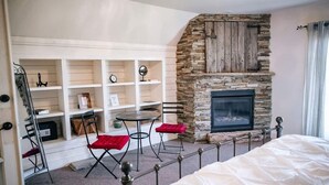 A bedroom with a stone fireplace, a bed, a small table with red cushions, and a metal railing. - Granary Two Bedroom Luxury Suite (Dallas)