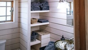 A compact bathroom with wooden slats on the ceiling, a built-in shelf with towels, a sink with a decorative bowl, and a hanging light fixture. - Granary Two Bedroom Luxury Suite (Dallas)