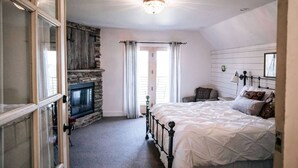 A bedroom with a stone fireplace, a bed with white bedding, a nightstand, and a window with curtains. - Granary Two Bedroom Luxury Suite (Dallas)